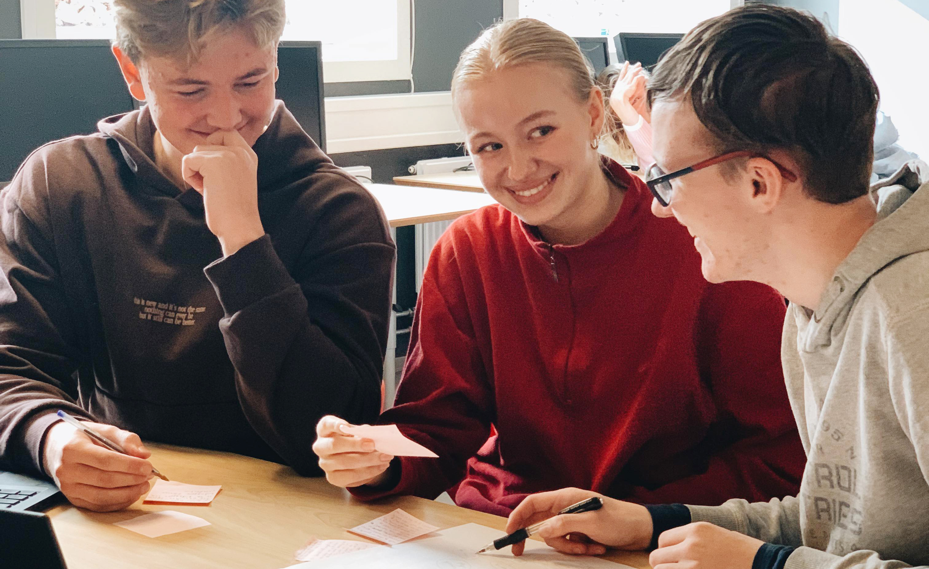 Tre ungdommer snakker sammen ved en pult. Inne.