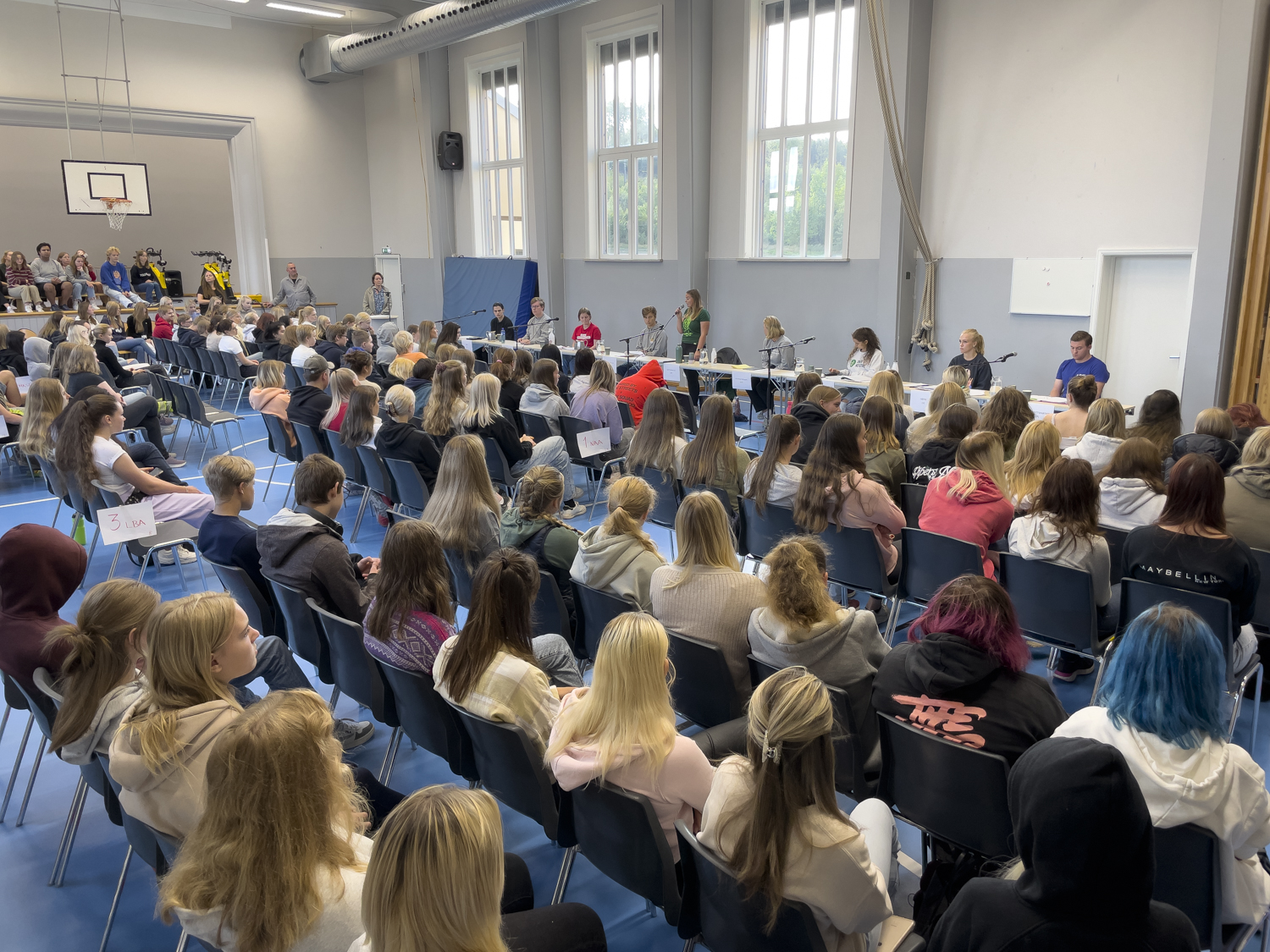 Gymsal fylt med elever som hører på 9 ungdomspolitikere som sitter i et panel.