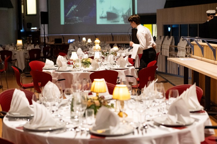 Runde bord med røde stoler rundt, dekket med hvite servietter, glass, bestikk og tallerkner.