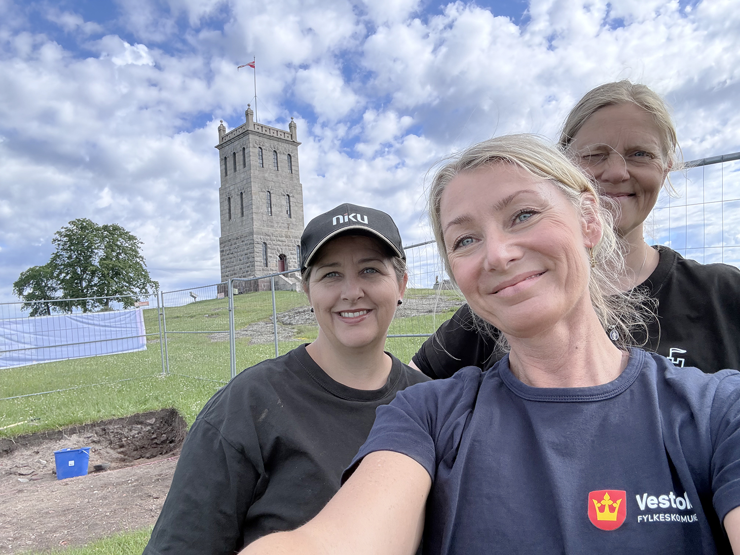 Tre blide damer foran tårnet på Slottsfjellet. Mellom dem og tårnet er området hvor de holder på å grave.