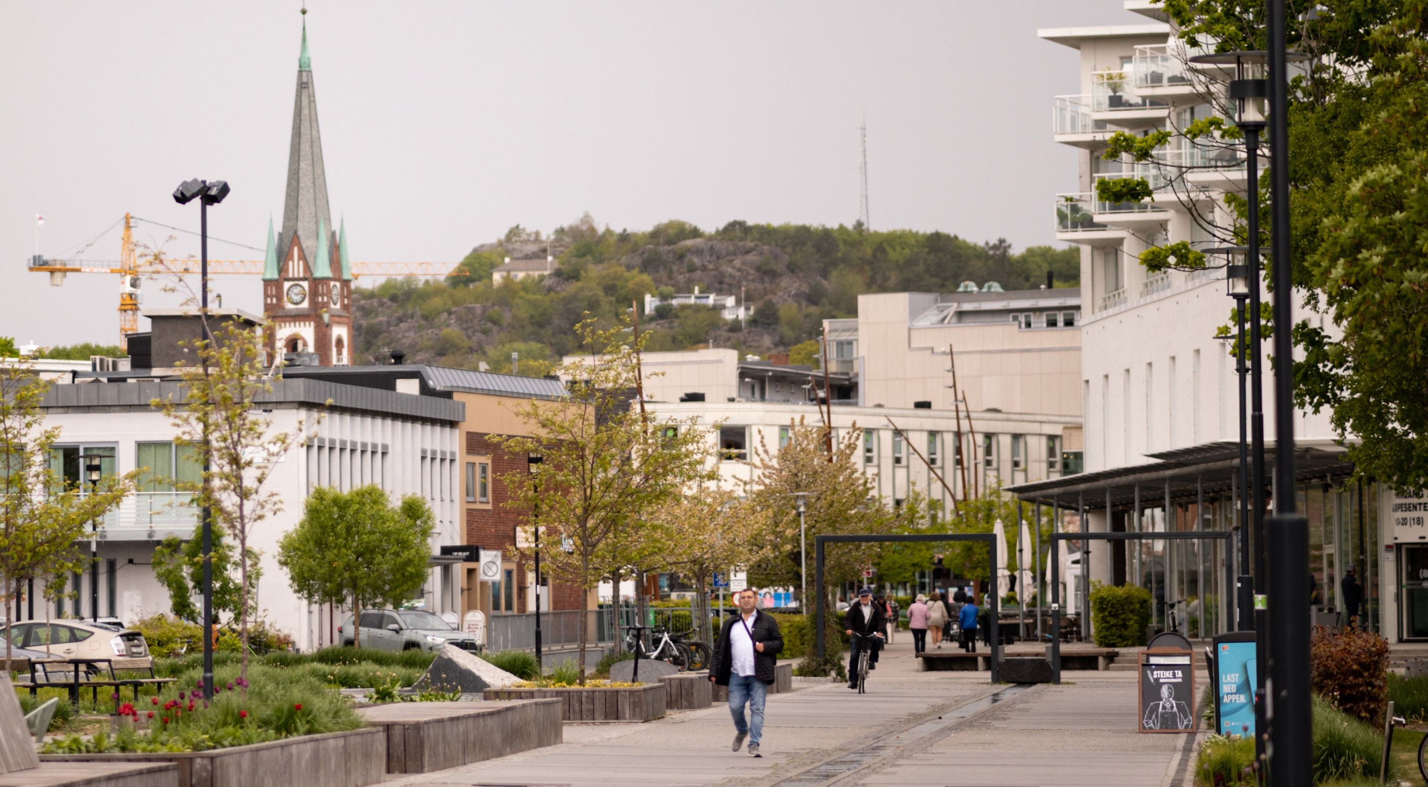 Sparsomt befolket gågate i bysentrum mellom grøntarealer under en overskyet himmel. Enkelte mennesker på vei til og fra steder ses på avstand. Nærmest kommer en mann i olabukser, hvit t-skjorte og cardigan mot oss med en veske på over skulderen. I bakgrunnen ser vi et kirkespir, en heisekran og en ås med noen villaer, noen steinknauser, trær og en mast. De som er kjent vil kunne se at det dreier seg om Sandefjord.