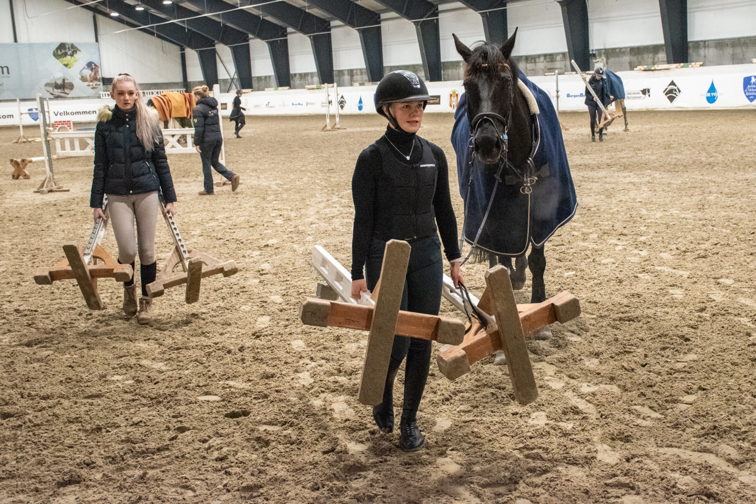 To elever bærer hinder i ridehallen. Den ene leier også en hest.