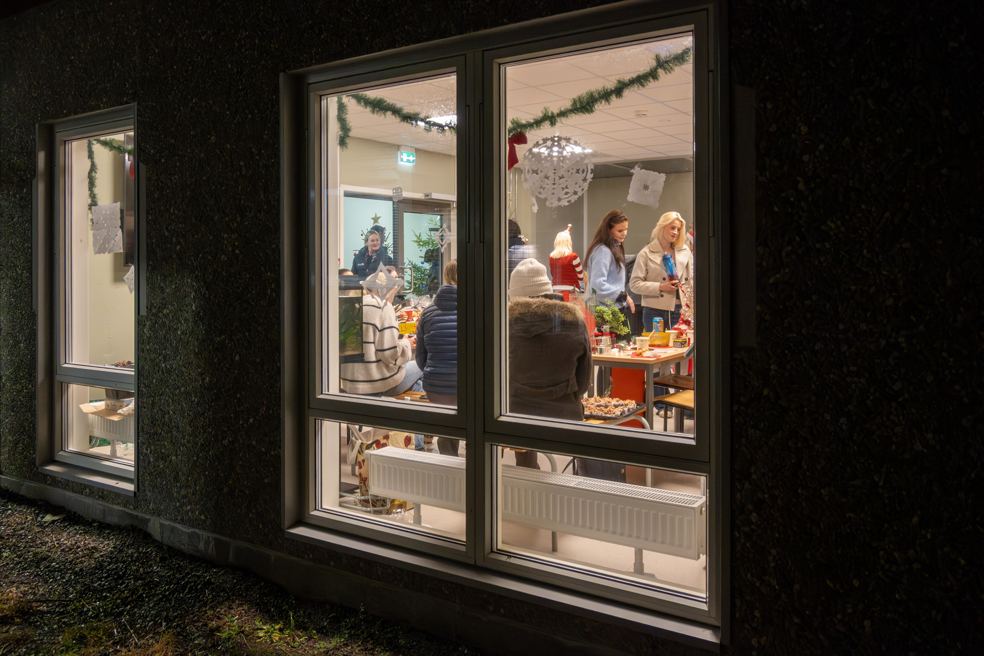 Vi ser inn gjennom vinduer. Mørkt ute. Lyst og mennesker inne.