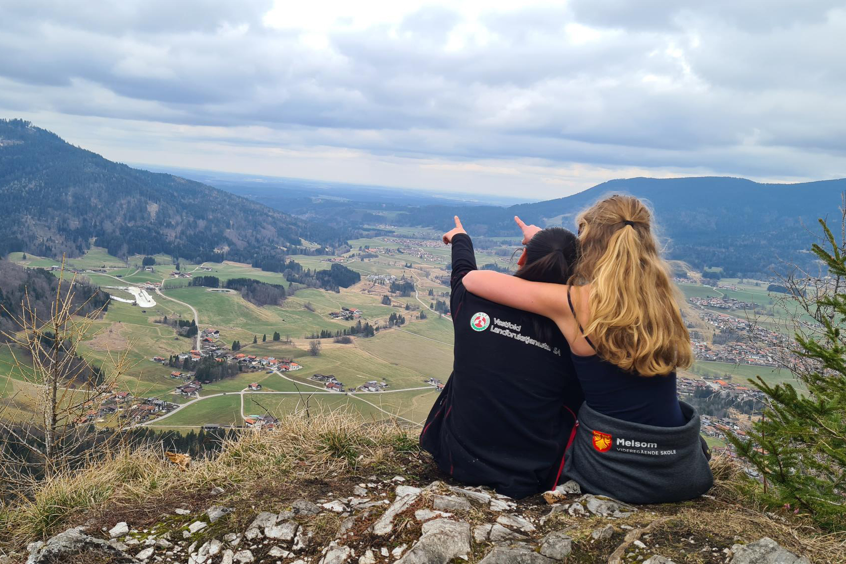 Ryggen til to jenter som sitter og ser ut over en dal