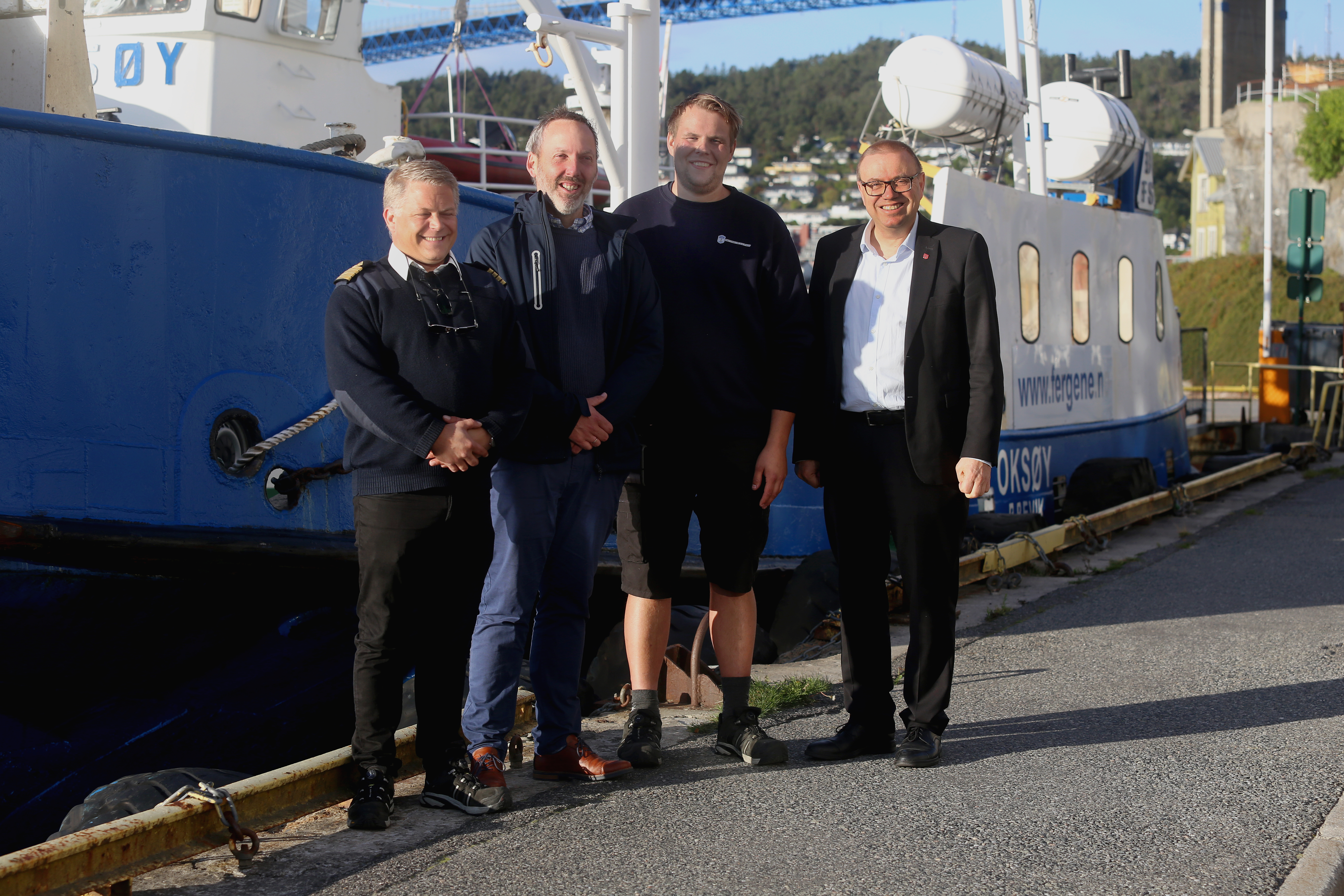 Fire menn er i fokus i midten av bildet. Bak skimtes ferja Oksøy.