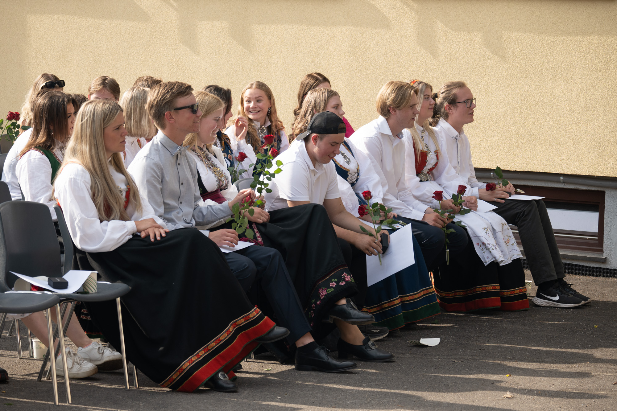 Gutter og jenter i pene klær sitter i stoler, ute.