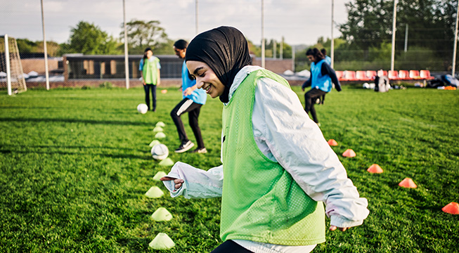 Flere barn i blå og hvite fotballdrakter fører ball mellom kjegler mens en voksen kledt i svarte treningsklær gir instruksjoner.