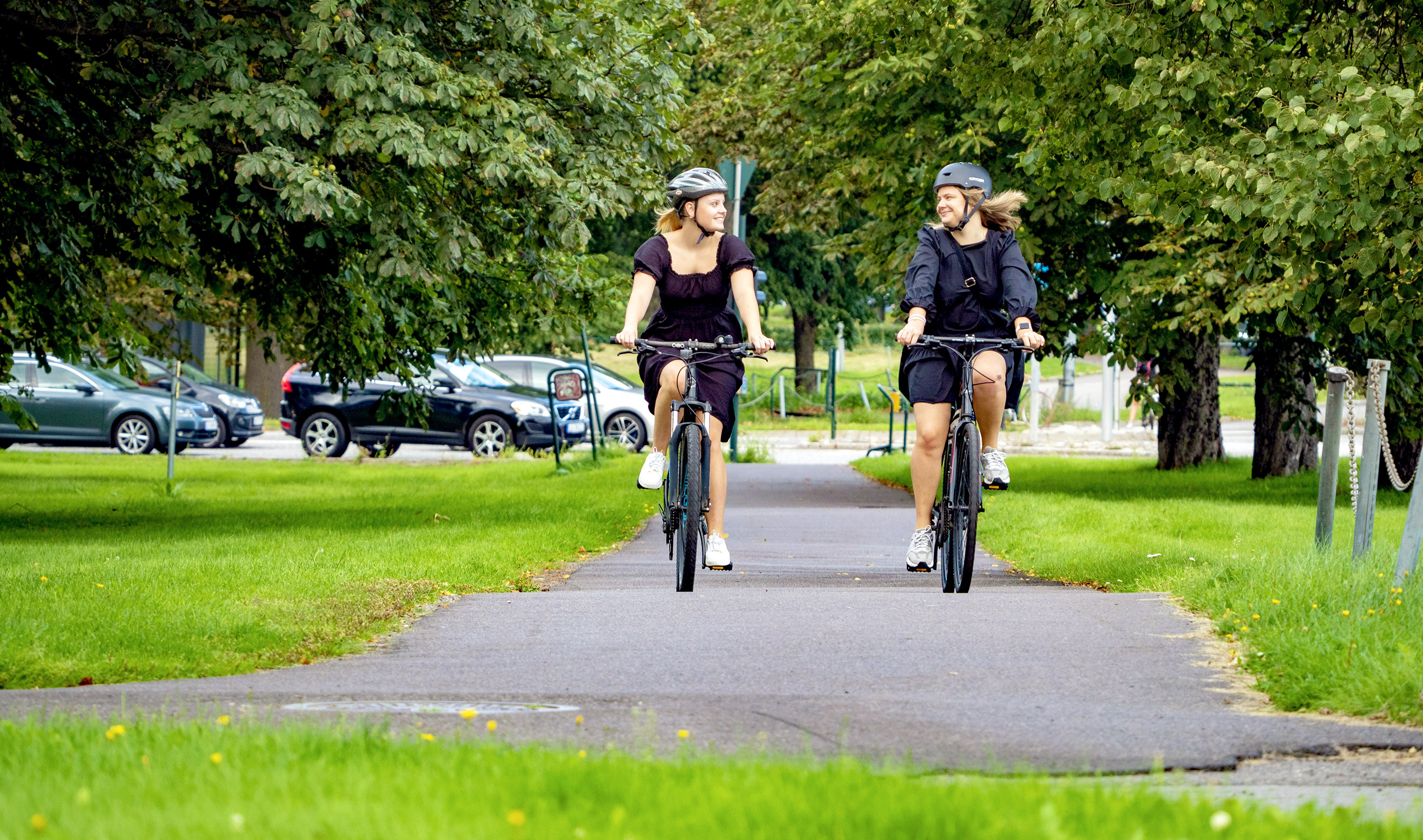 To kvinner sykler på sykkelvei ved Mammutkrysset omkranset av store grønne trær